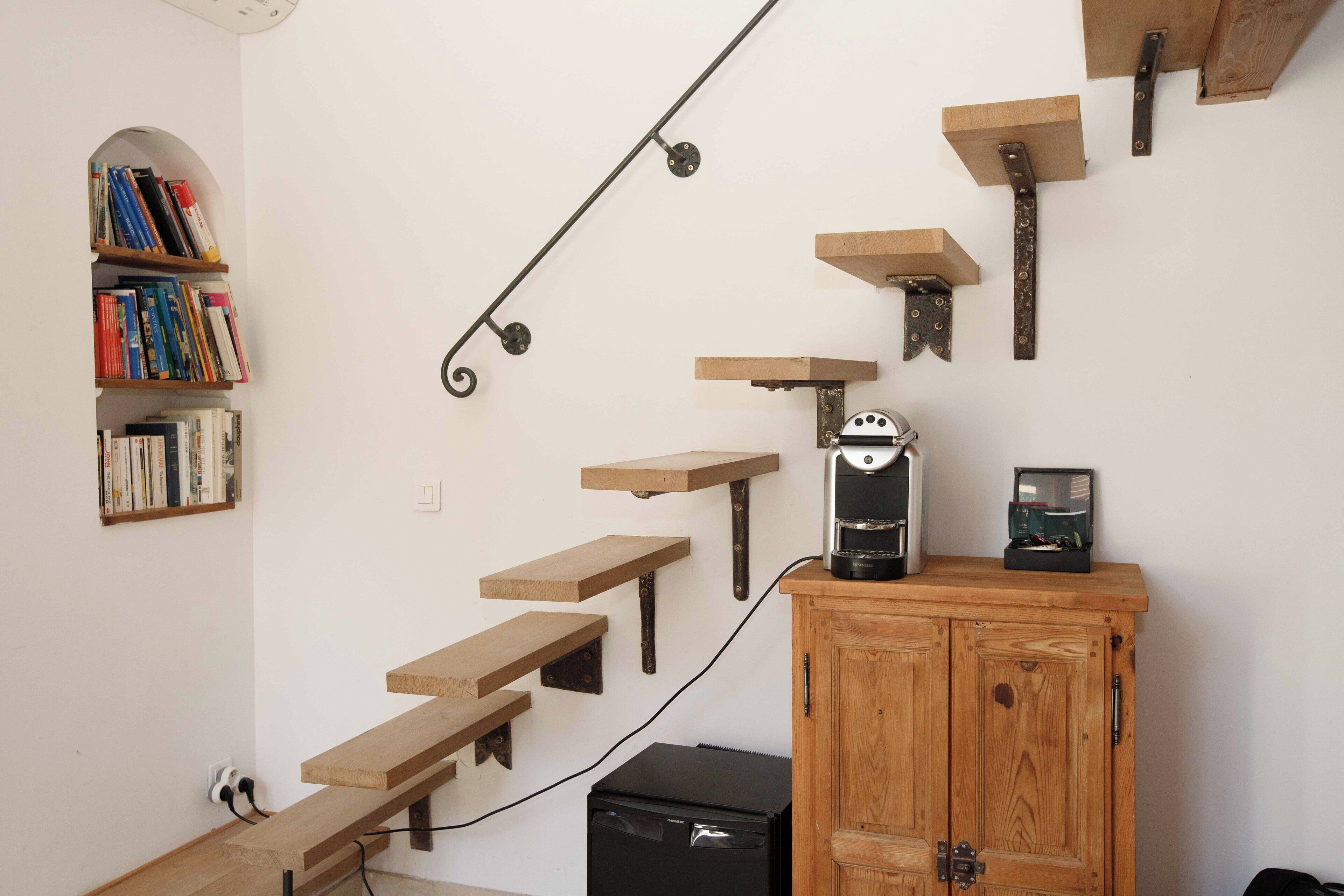 A staircase with wooden steps and metal brackets, a coffee maker on a wooden shelf, and a bookshelf with books.