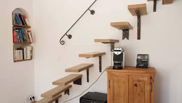 A staircase with wooden steps and metal brackets, a coffee maker on a wooden shelf, and a bookshelf with books.