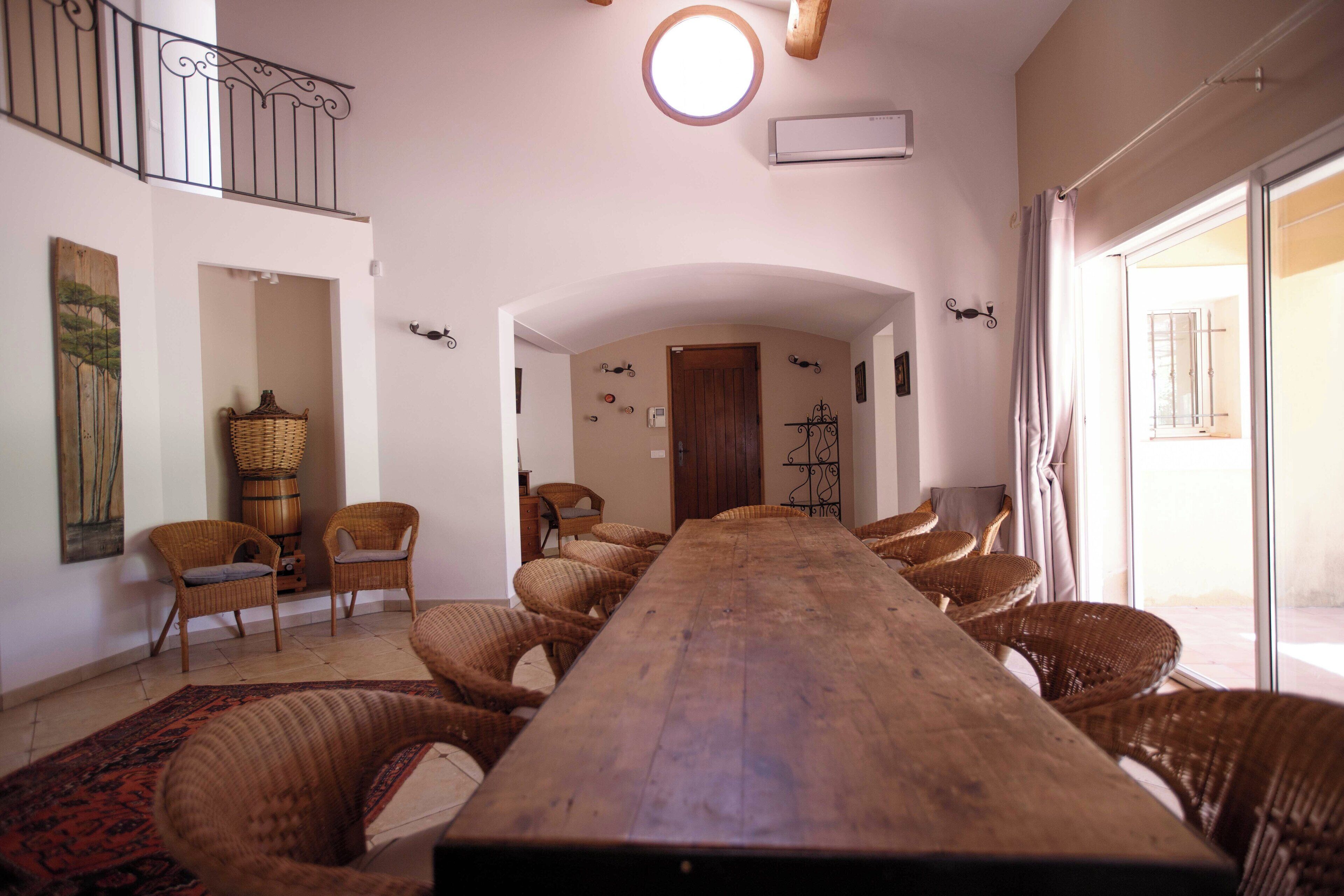Een lange houten tafel met rieten stoelen, een grote geweven mand en een houten vat in een lichte kamer met witte muren en een balkon.