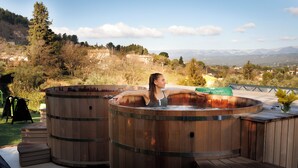 Iemand ontspant in een houten jacuzzi met uitzicht op een bergachtig landschap.