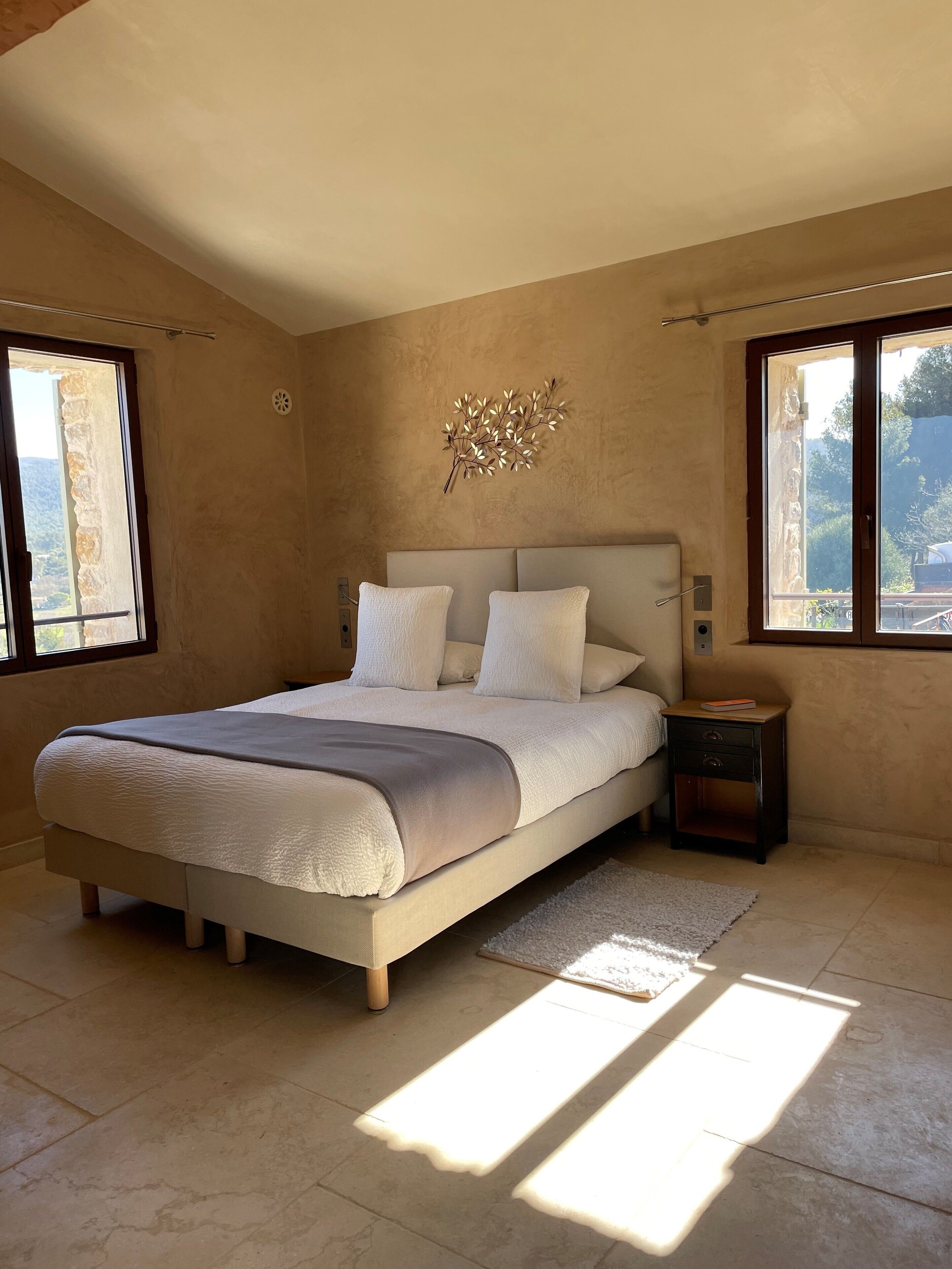A bedroom with a large bed, a nightstand, a wall art piece, and a view of the outdoors through the windows.