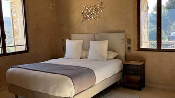 A bedroom with a large bed, a nightstand, a wall art piece, and a view of the outdoors through the windows.
