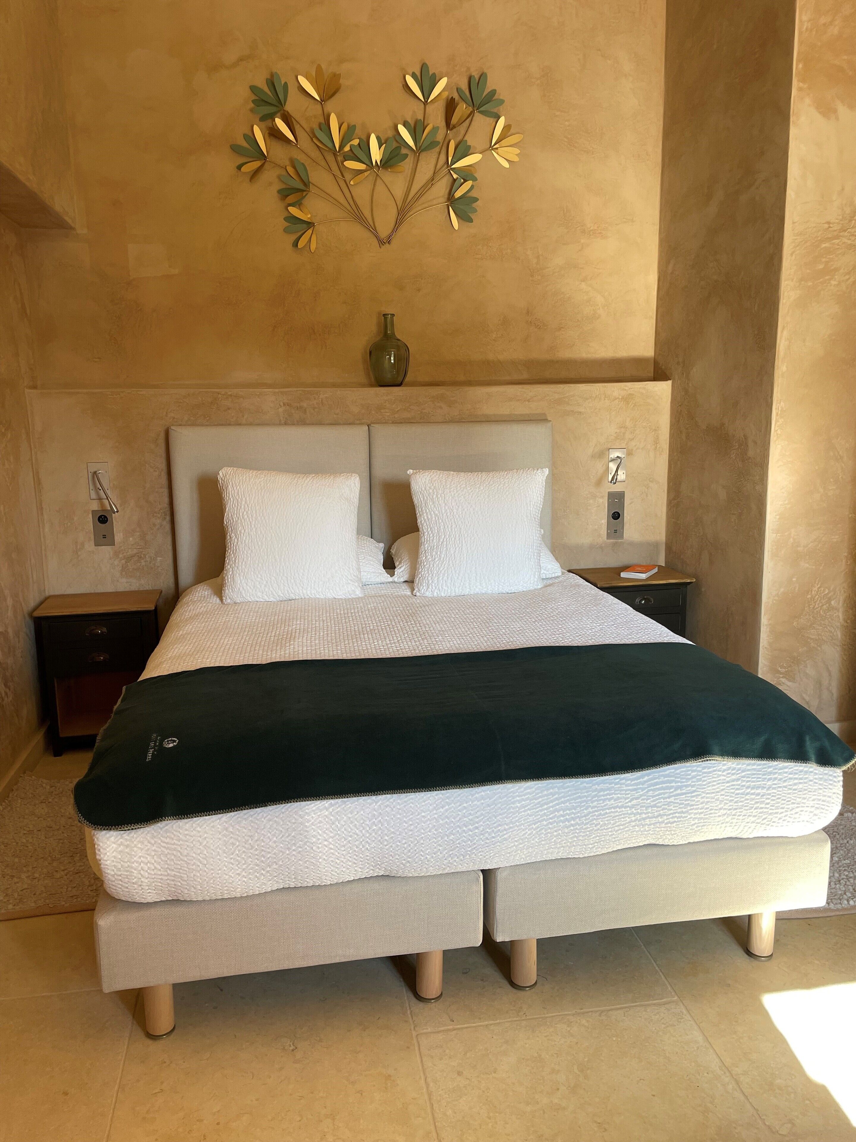 Un lit double avec une couverture blanche et verte, deux oreillers blancs, une tête de lit beige, une table de chevet avec une lampe et un élément décoratif fixé au mur.