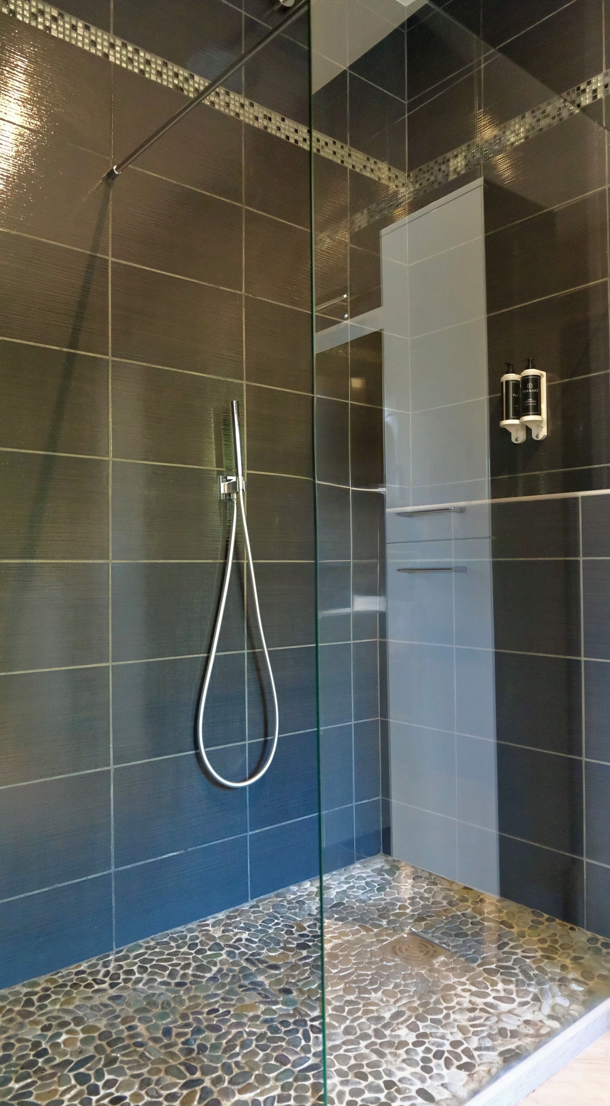 Ducha con paredes de azulejos, piso con textura de guijarros y ducha de mano.