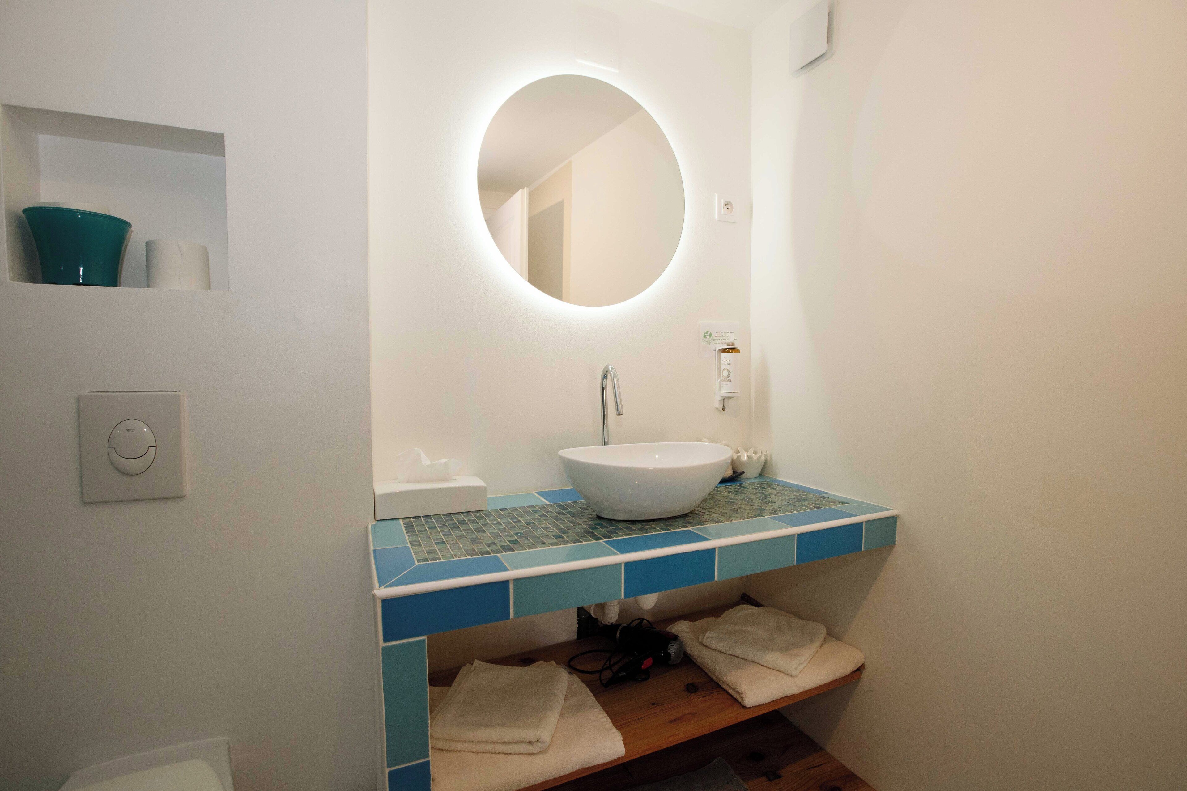 A bathroom with a white sink, blue tiles, a round mirror, and a shelf with towels.