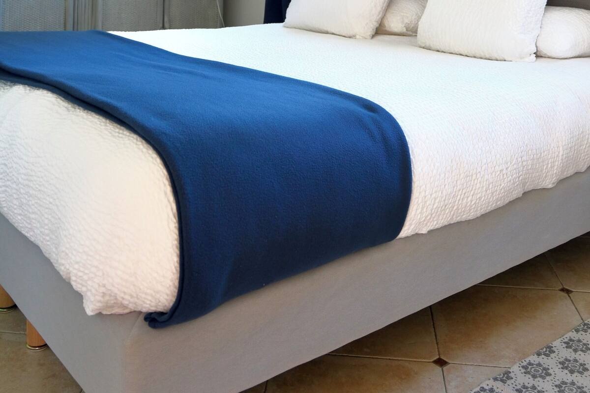 Un dormitorio con cama, ventana con cortinas, lámpara, cuadro enmarcado en la pared y una mesita con lámpara.