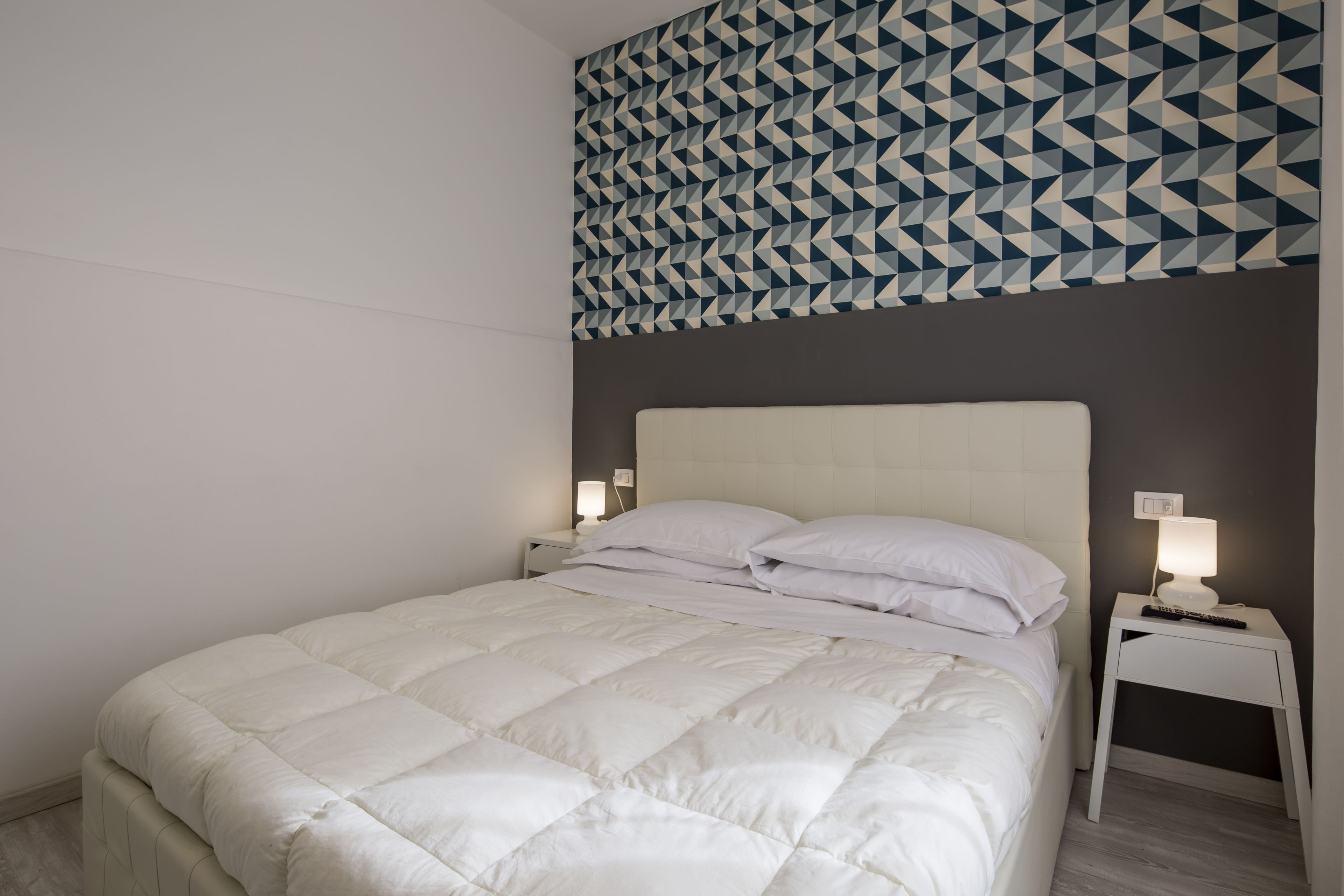 Een netjes opgemaakt bed met een witte dekbedovertrek, twee nachtkastjes met lampen, en een geometrisch patroon op de muur boven het bed.