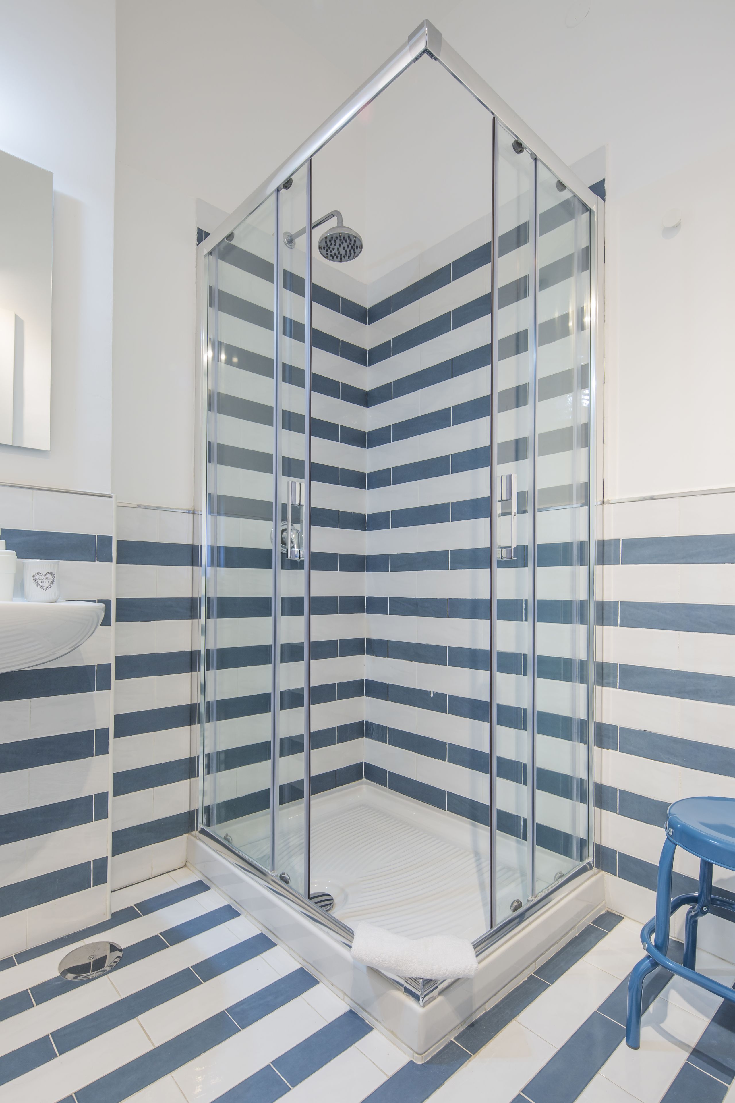Une salle de bain moderne avec une douche à parois de verre, des carreaux à rayures et un tabouret bleu.
