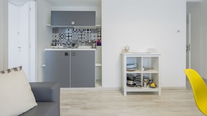 Un espacio habitable pequeño con kitchenette, un sofá gris, un estante blanco con utensilios de cocina y una silla amarilla.
