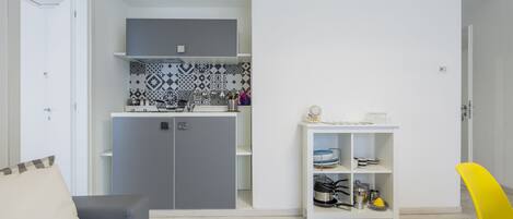 Een ruimtelijk ingerichte leefruimte met kitchenette, een grijze bank, een witte plank met keukenspullen en een gele stoel.