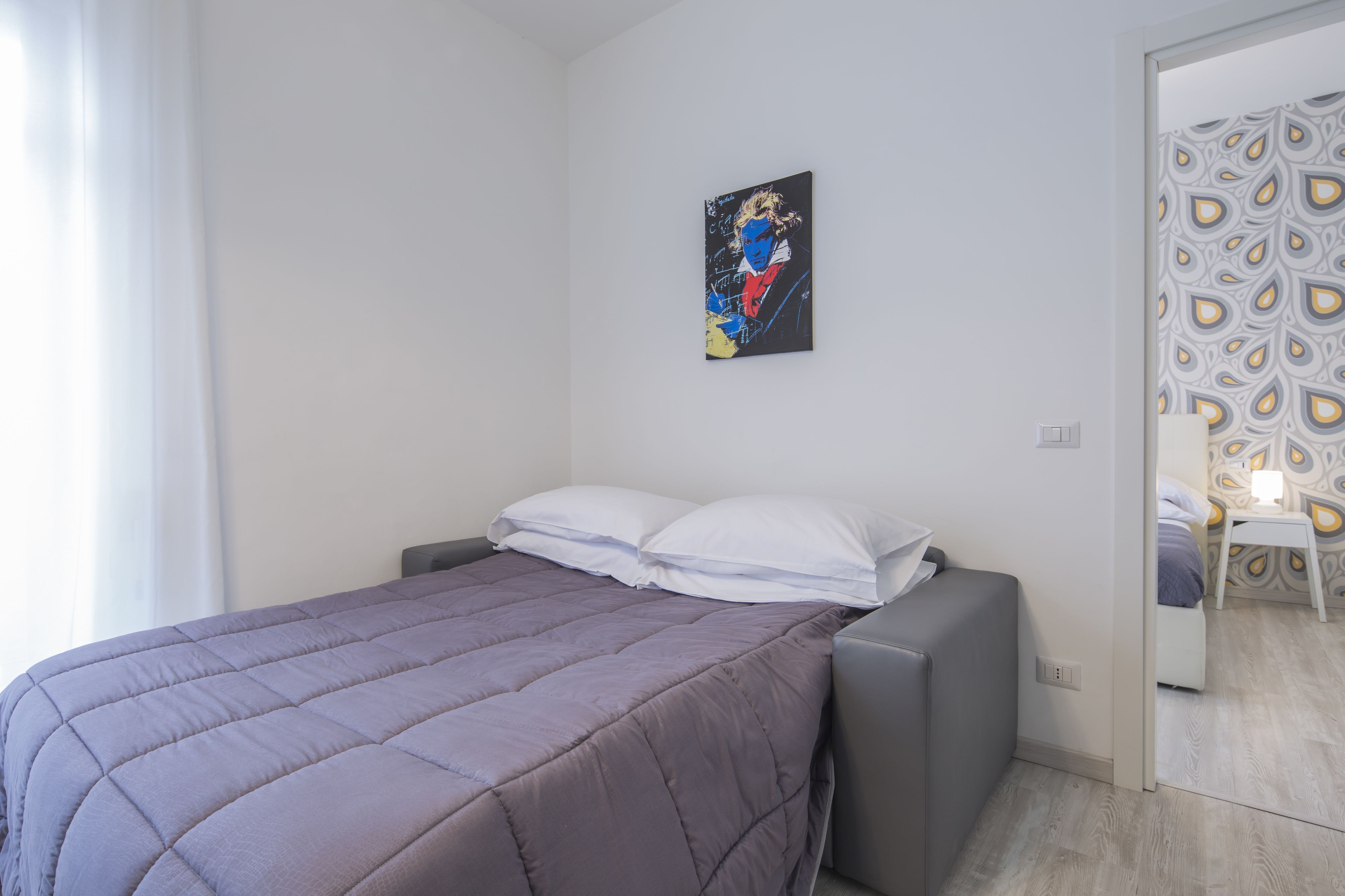 A bedroom with a bed, a gray sofa, a wall-mounted artwork, and a patterned wallpaper in the adjacent room.