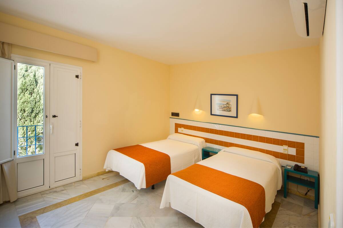 Une chambre d’hôtel avec deux lits, une fenêtre, une porte et une petite table avec un téléphone.