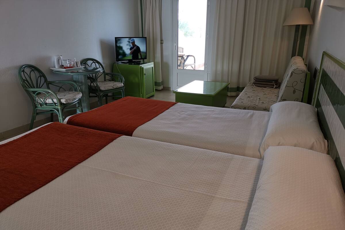Une chambre d’hôtel avec deux lits, une télévision, un bureau vert et une fenêtre avec des rideaux.