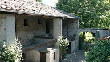A rustic stone building with a sloped roof, a small porch, and a gravel pathway leading to a wooden bridge.