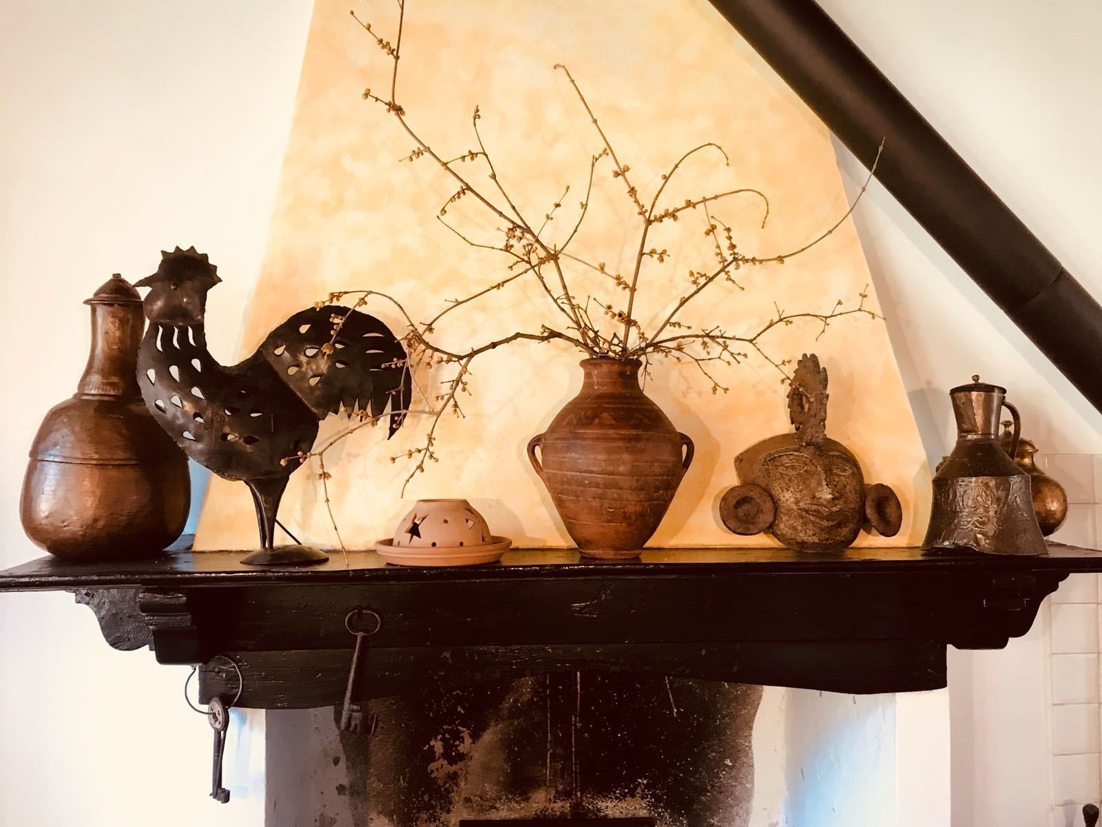 Una mensola rustica con diversi oggetti d'antiquariato, tra cui un vaso in ceramica, una scultura di un gallo in metallo e una piccola pianta in vaso.