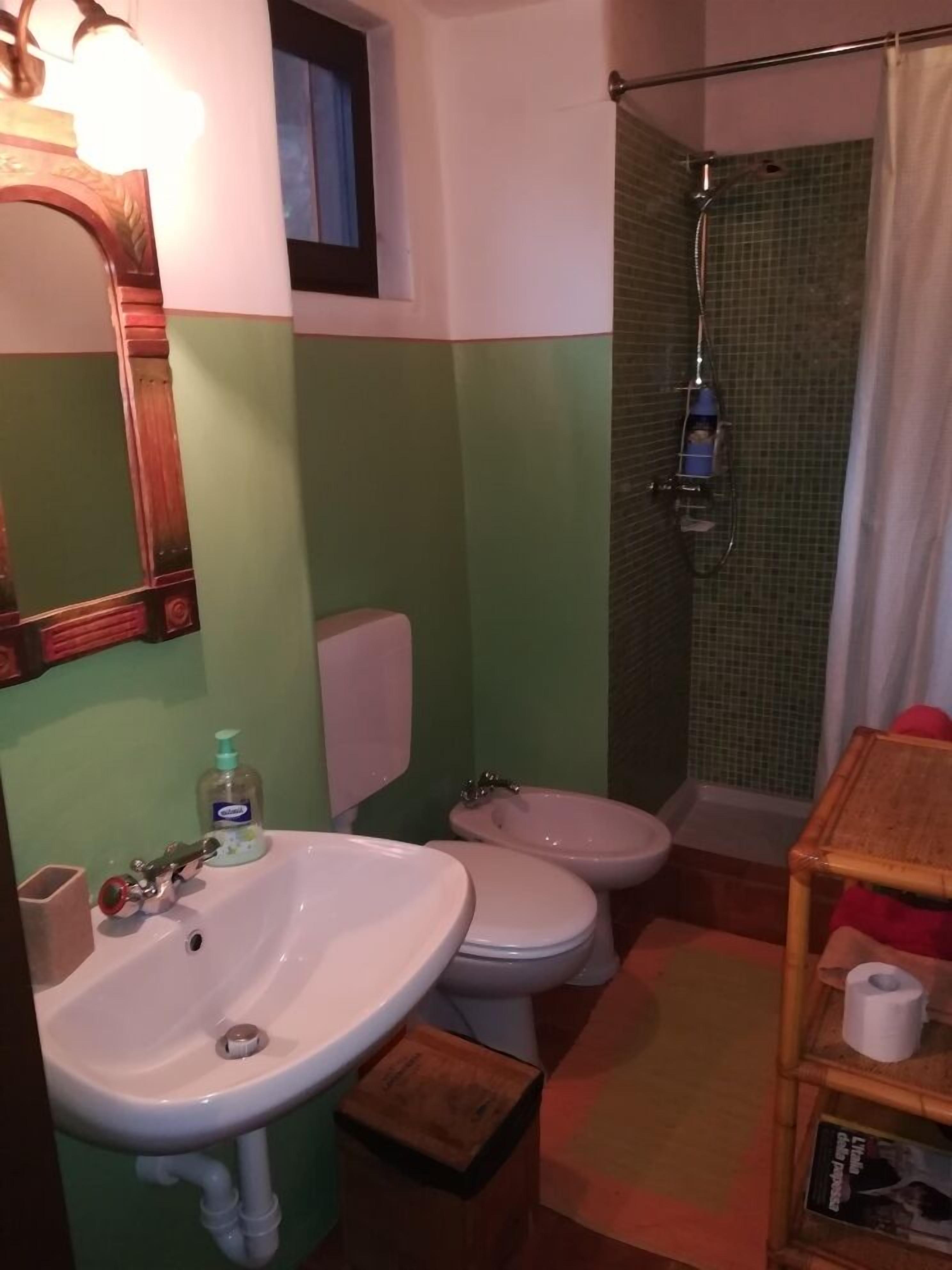 A bathroom with a sink, two toilets, a shower, and a wooden shelf with a toilet paper holder.