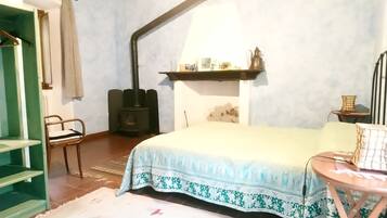 Un dormitorio con una cama, una silla, una mesita, un estante con objetos y una estufa negra.