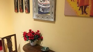 A room with a wooden chair, a small table with a vase of red flowers, a framed picture of a coliseum, and a painting of people in orange robes. - La Giara Rooms (Verona)