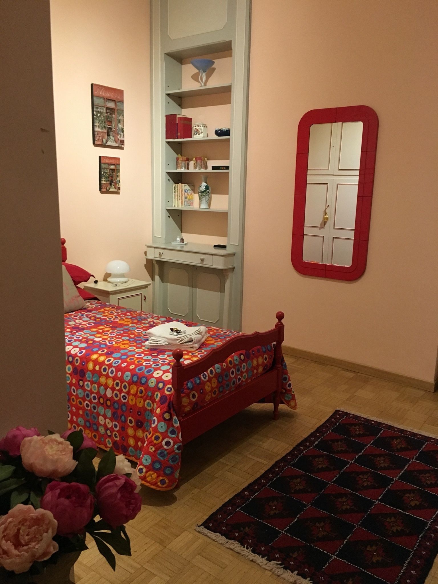 A bedroom with a bed, a nightstand, a dresser, a mirror, and a small shelf with books and decorative items.