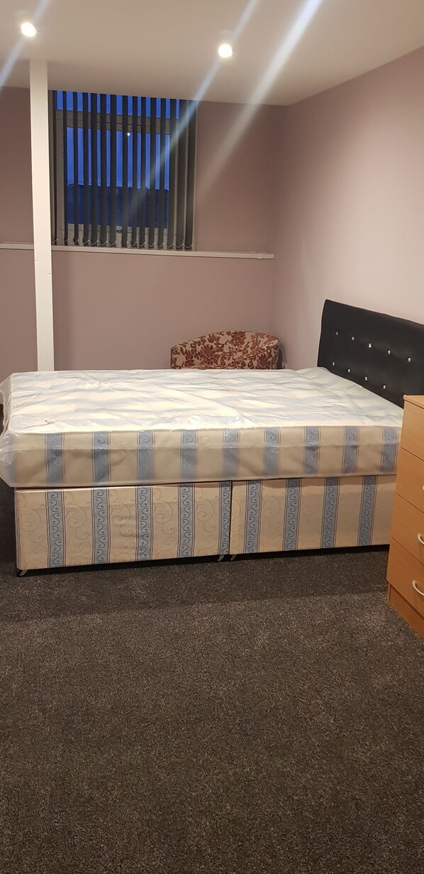 Une chambre à coucher avec un lit, une petite banquette, une tête de lit et une fenêtre avec des stores.