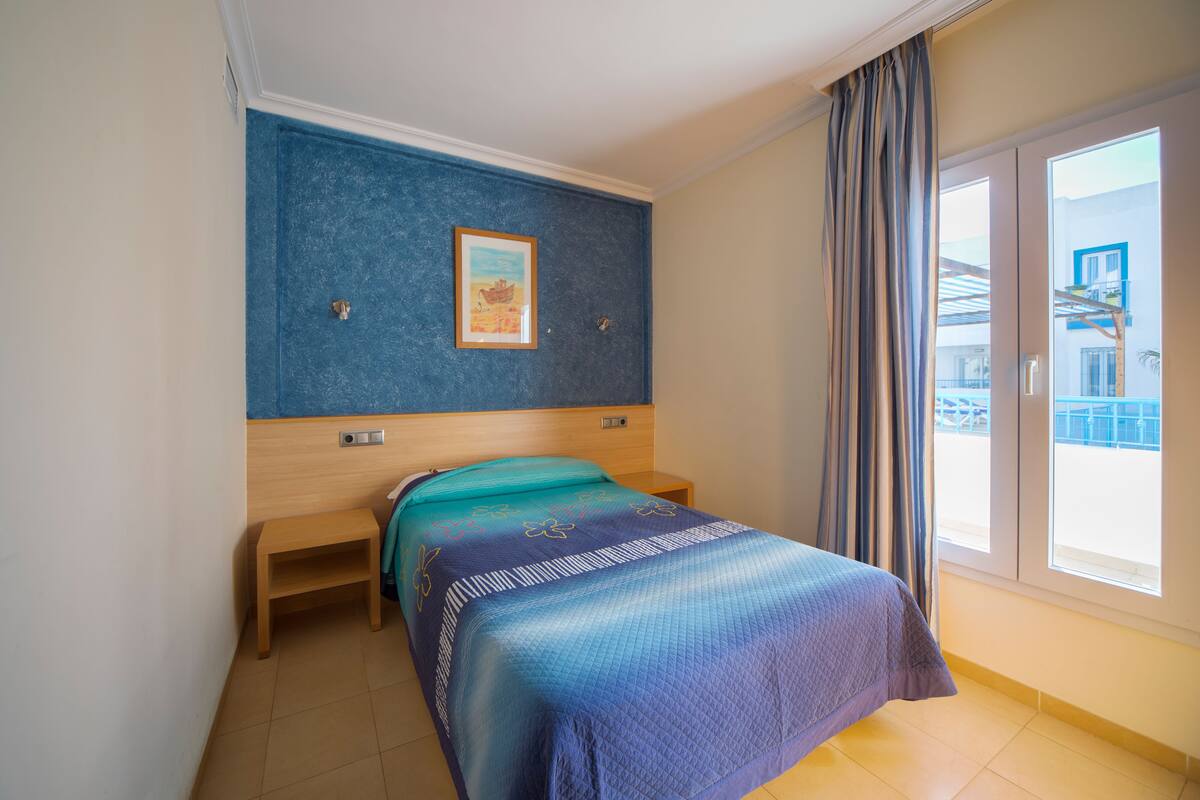 Une chambre à coucher comprenant un lit, un petit banc en bois, une table de chevet, une étagère fixée au mur et une fenêtre avec des rideaux.