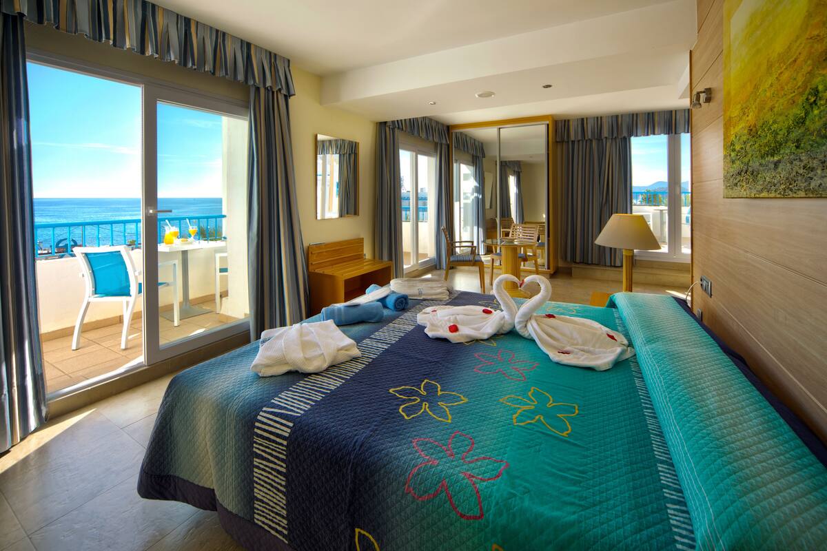 Une chambre d’hôtel avec un balcon, un lit recouvert d’une couvre-lit à motifs floraux, un bureau avec une chaise et une vue sur la mer.