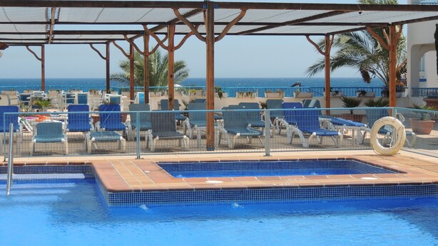 A pool area with blue lounge chairs, a tiled pool, and a view of the sea. - Hotel Punta del Cantal  (Mojacar)
