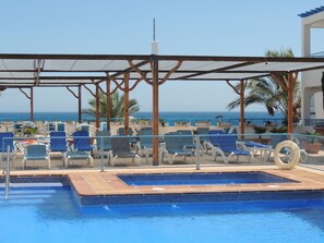 Zona de piscina con sillas reclinables azules, una piscina con baldosas y vistas al mar.