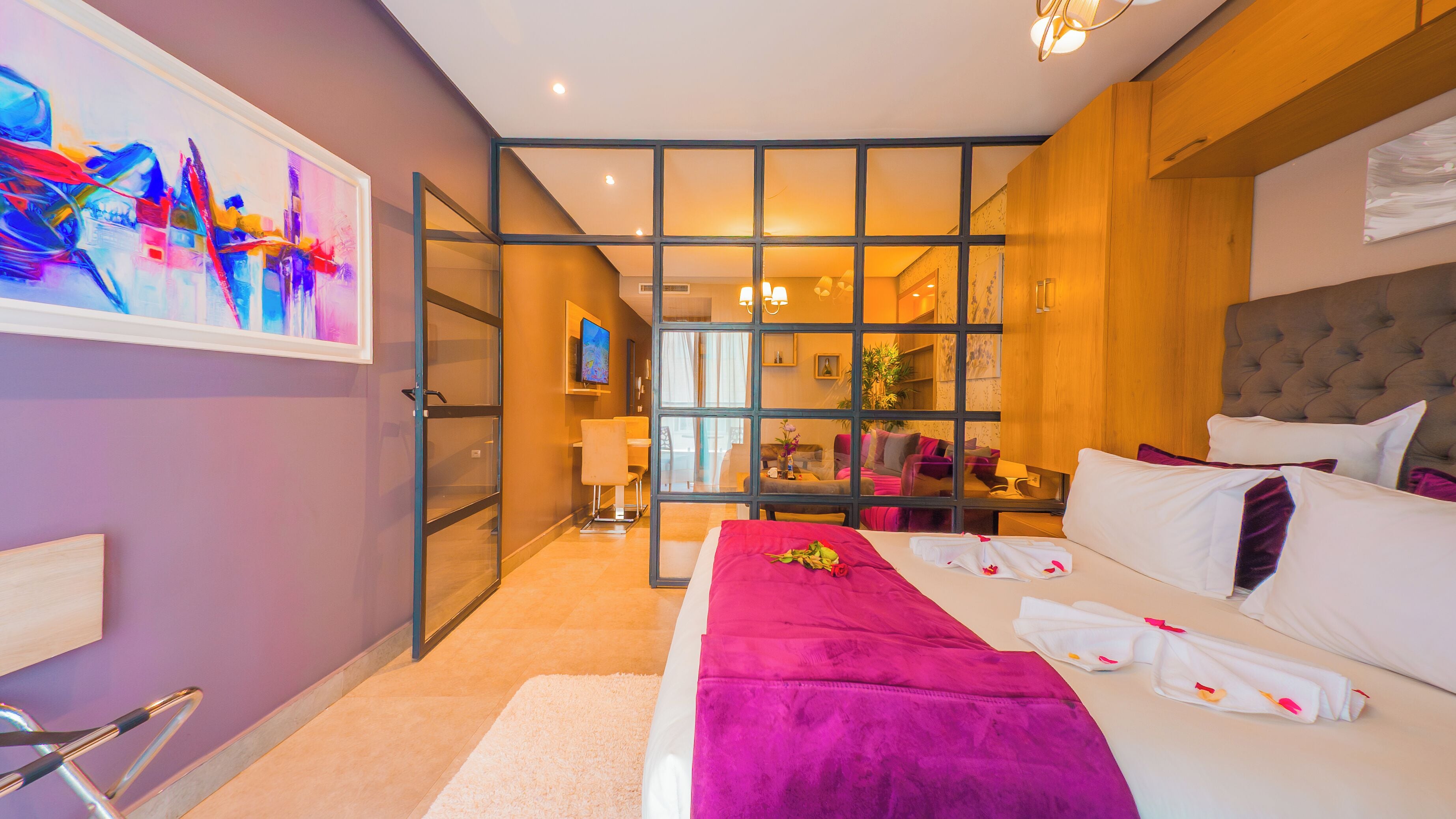 A modern hotel room with a large bed, a glass partition, and a view of the living area.
