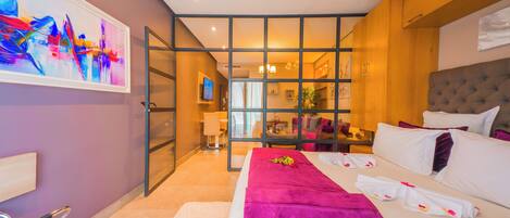 A modern hotel room with a large bed, a glass partition, and a view of the living area.