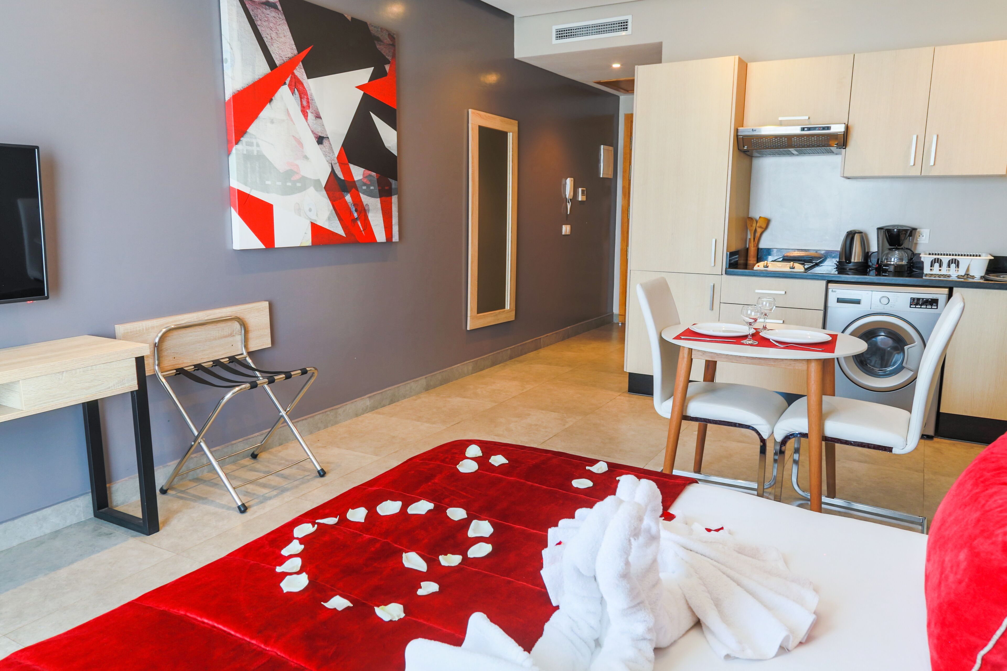 A compact hotel room with a bed, a red rose petal carpet, a desk with a chair, a washing machine, and a kitchenette.