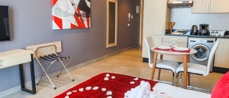 A compact hotel room with a bed, a red rose petal carpet, a desk with a chair, a washing machine, and a kitchenette.