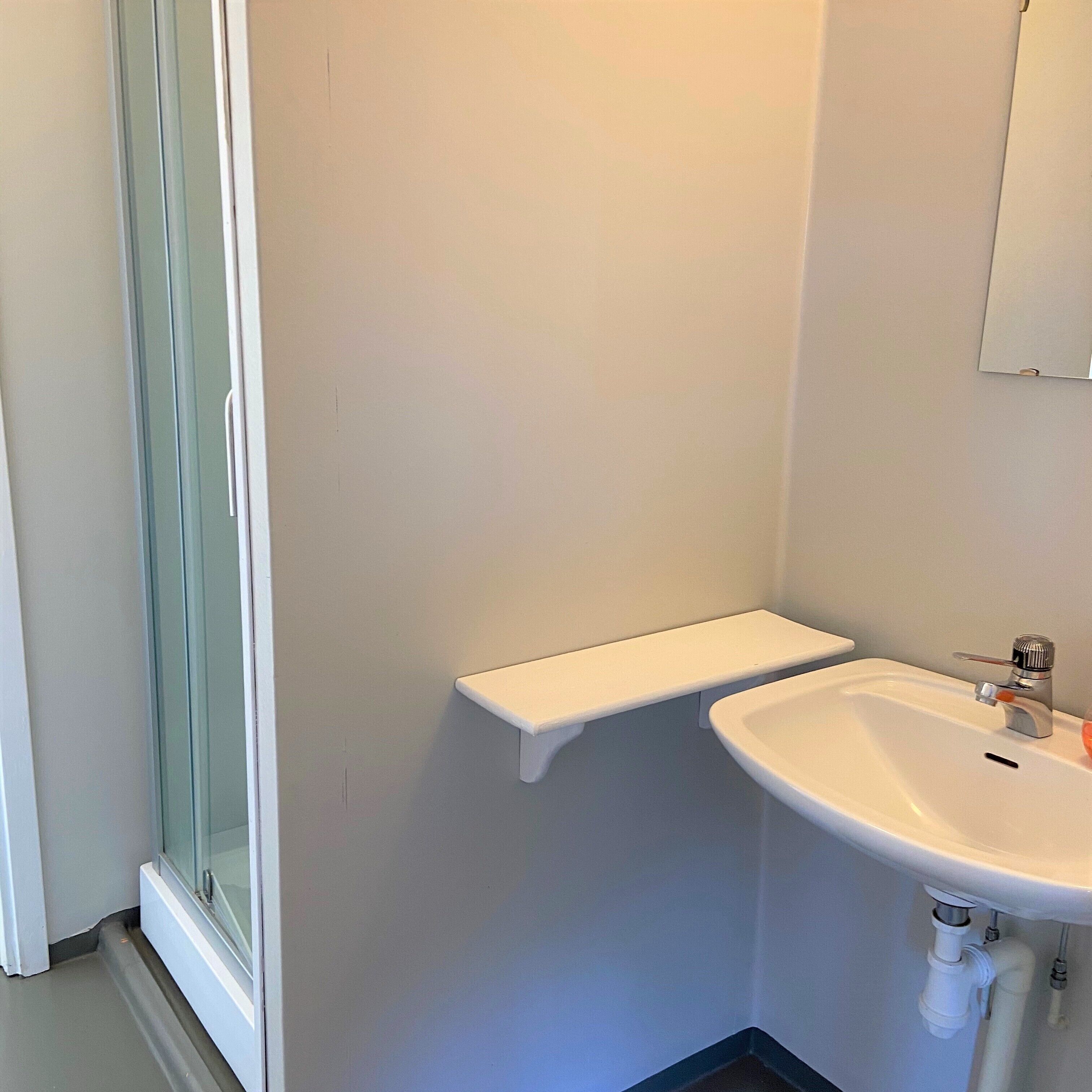Een badkamer met een douchecabine van glas, een witte wastafel en een klein wit plankje.