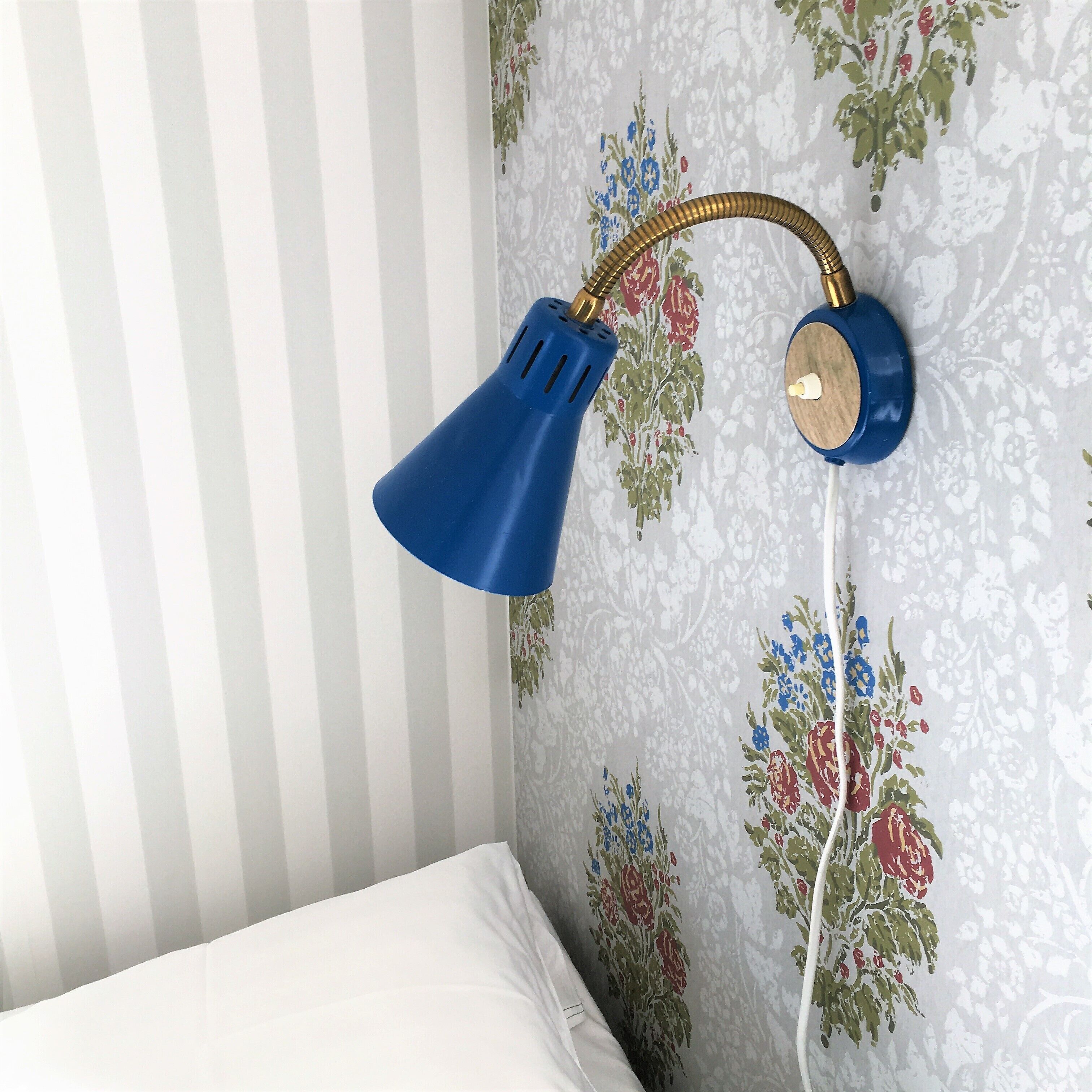 Un llit amb roba de llit blanca, una làmpada blava penjada a la paret i paper pintat amb motius florals.