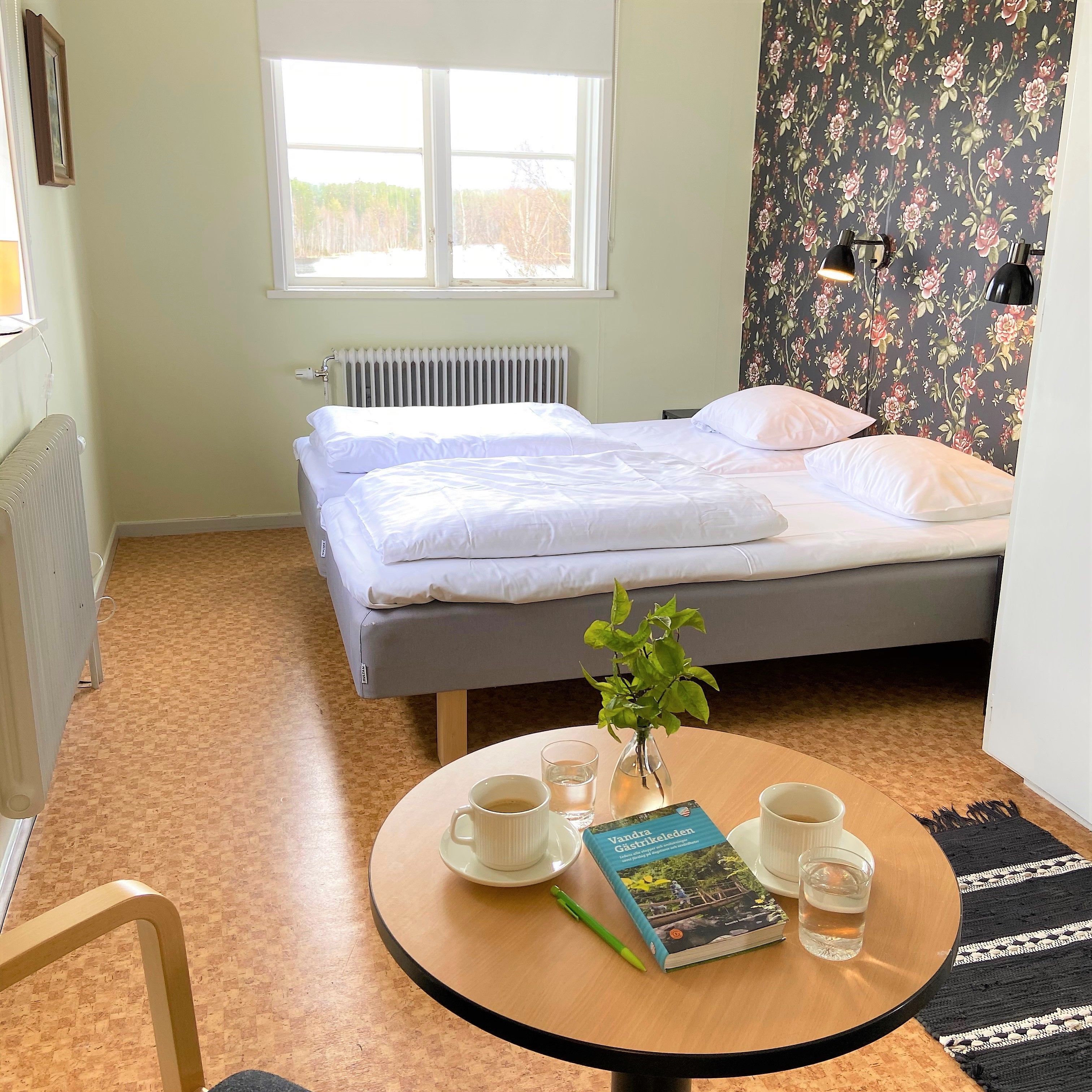 Un dormitorio con cama, un radiador, una ventana, una mesa con un libro, vasos y una planta.