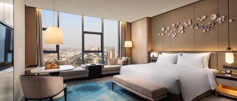 A modern hotel room with a large bed, a seating area with a view, and a cityscape visible through the windows.