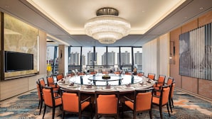 Une salle de conférence moderne avec une table ronde, des chaises orange, un lustre, un grand téléviseur et une vue sur la ville.