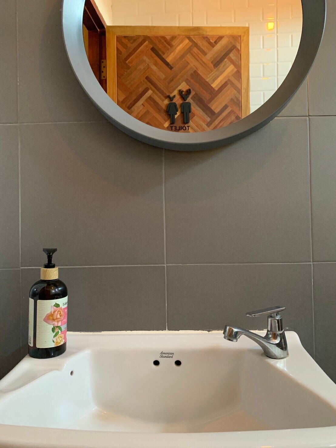 Un baño con un lavamanos blanco, un espejo redondo y una botella de loción.