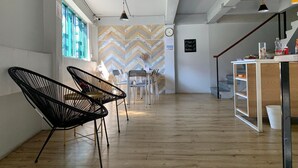 A modern interior with a herringbone-patterned wall, a wooden floor, black wireframe chairs, a white table, and a staircase with a wooden railing. - Busket Hostel (Chiang Rai)
