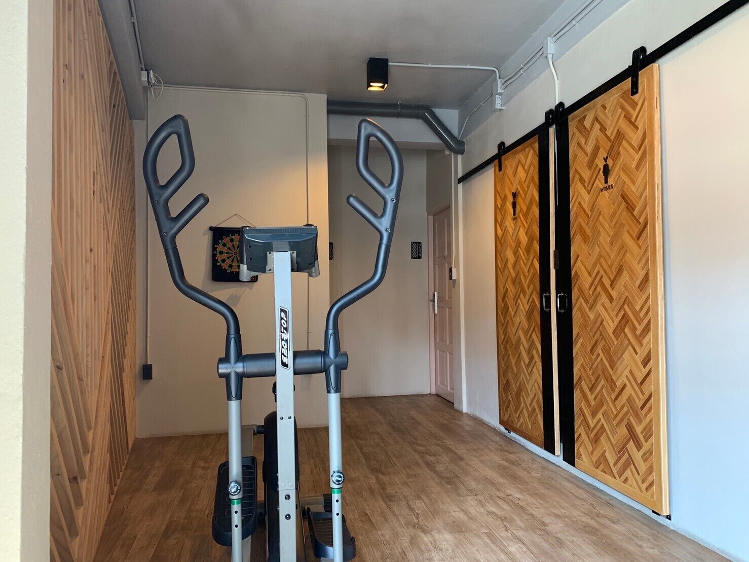 Ein Zimmer mit einem stehenden Ergometer, einer Dartscheibe und einer Schiebetür mit Holz-Fischgrätmuster.