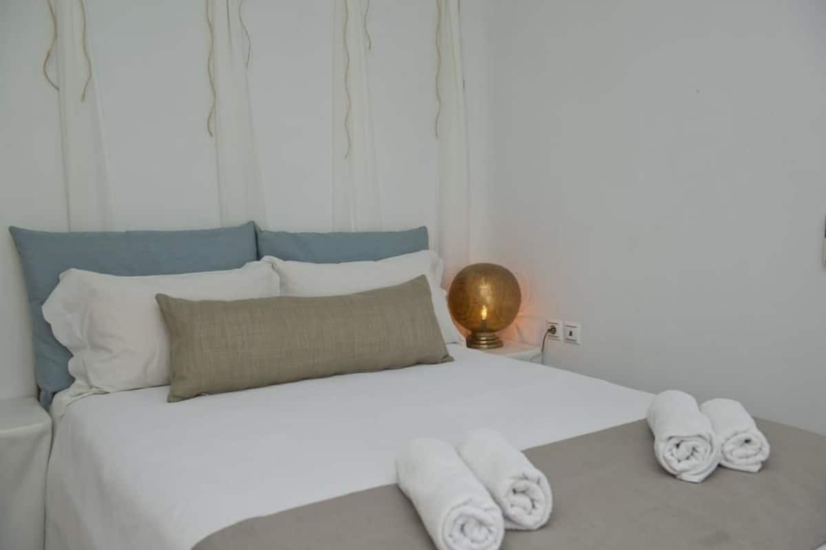 Een net opgemaakt bed met wit- en blauw beddengoed, een beige kussen en twee opgerolde handdoeken op het bed.