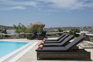 A poolside area with lounge chairs and a thatched umbrella, overlooking a coastal town. - Tatiana Villa & Apartments (Mykonos)