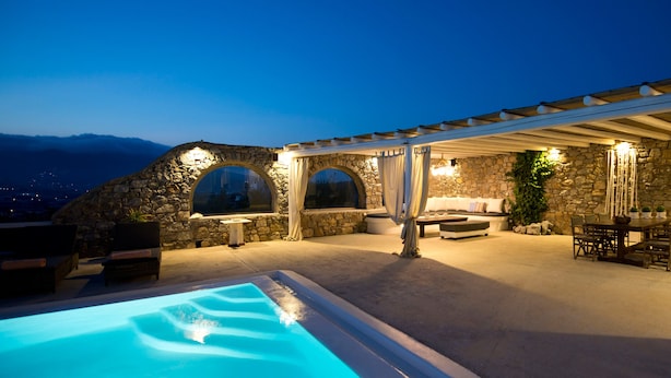 A poolside area with a stone wall, outdoor seating, and a covered patio. - Tatiana Villa & Apartments (Mykonos)