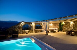 A poolside area with a stone wall, outdoor seating, and a covered patio. - Tatiana Villa & Apartments (Mykonos)