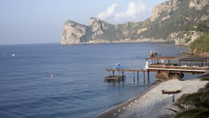 A coastal scene with a rocky cliff, a wooden pier extending into the sea, and a small boat on the shore. - Taverna del Capitano (Massa Lubrense)