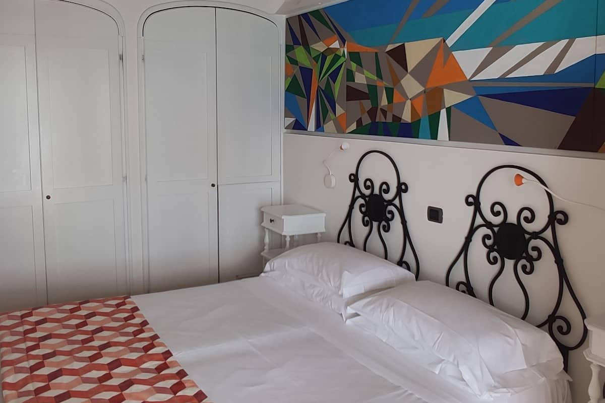 Un dormitorio con una cama, una mesita de noche blanca, una lámpara de pared y un edredón estampado con un motivo geométrico.