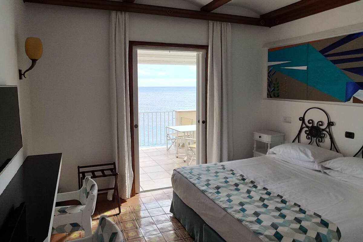 Un dormitorio con cama, escritorio, silla, un cuadro y vista al mar.