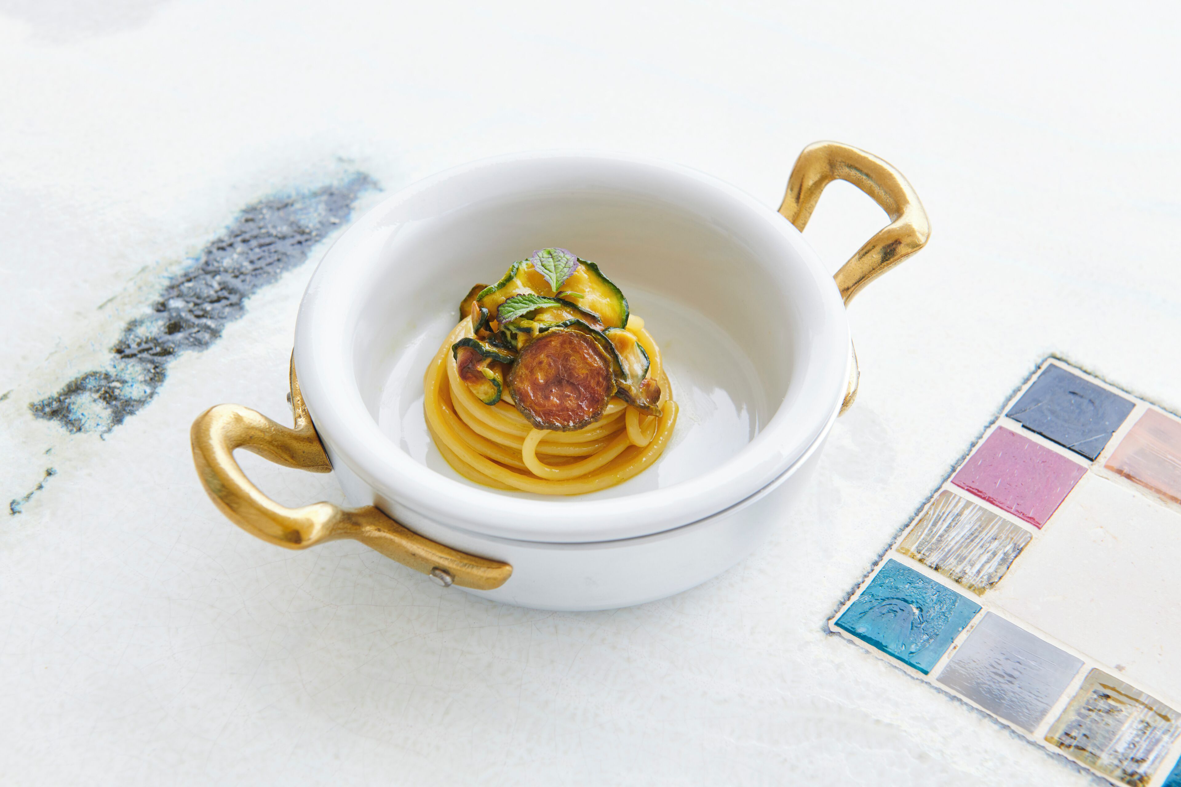 A white bowl with gold handles containing a pasta dish.