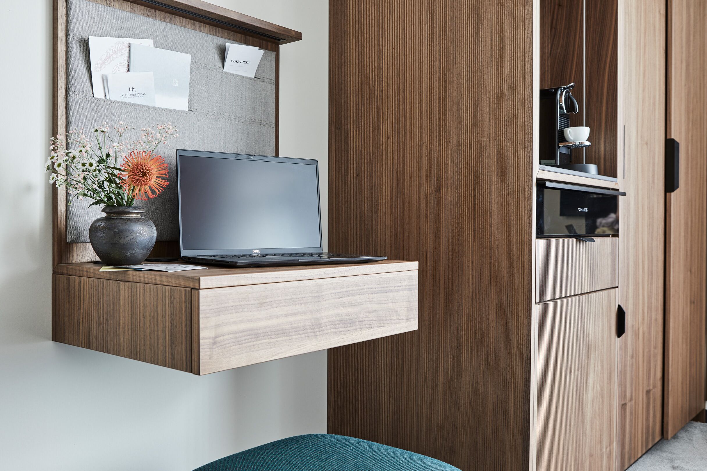 A modern kitchen with a built-in laptop on a wooden counter, a vase with flowers, and a coffee machine.