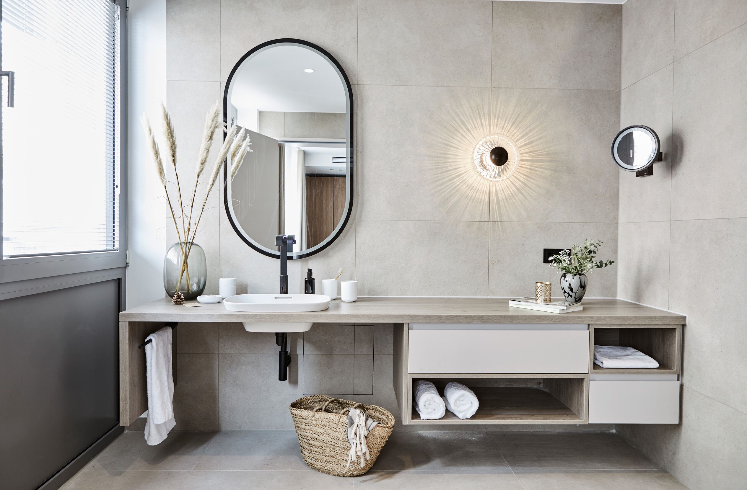 A modern bathroom with a large mirror, a round wall light, and a built-in shelf with towels.